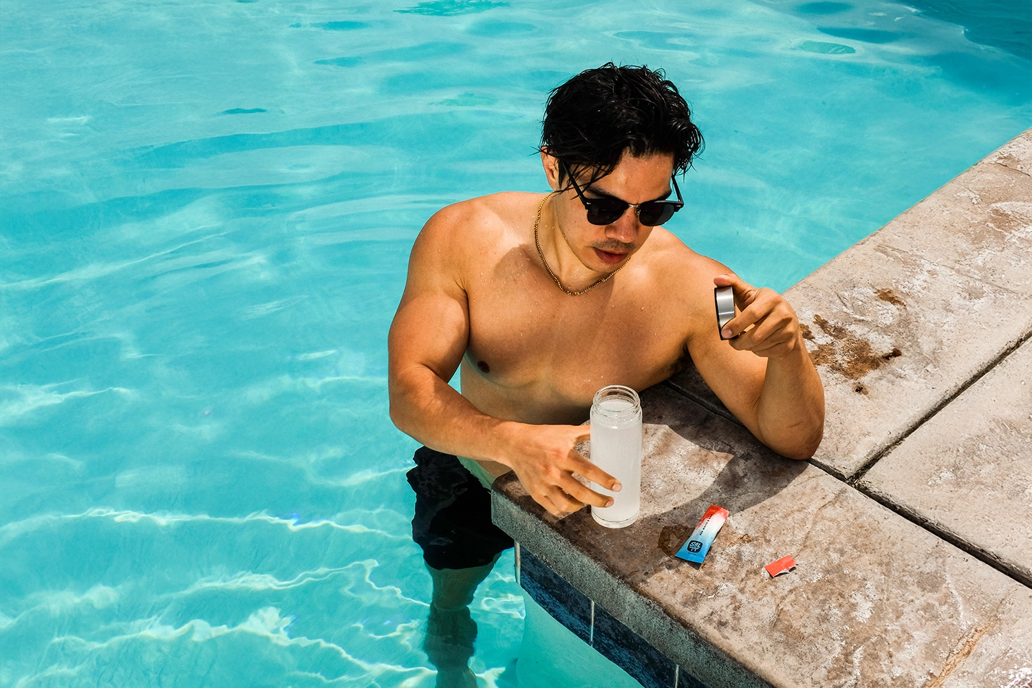 Man by a pool holding a water bottle of SALTT and a straw, with sunglasses on.