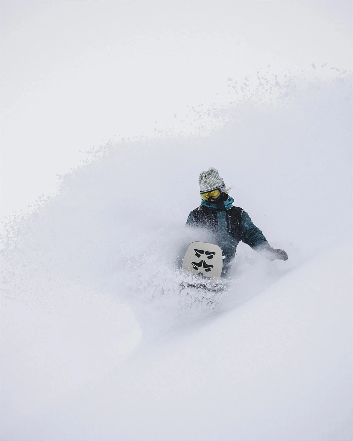 A light-colored snowboard with a black, abstract logo is ridden by a snowboarder through deep snow, creating a large white spray.
