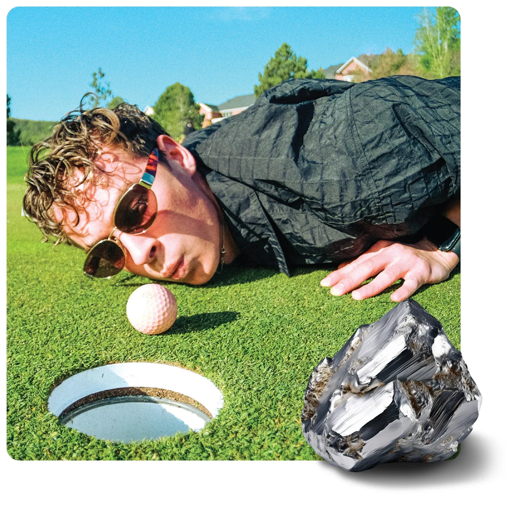 A man in sunglasses lies on a golf course green, looking at a pink golf ball by the hole. A large, dark, faceted metallic rock rests on the grass.