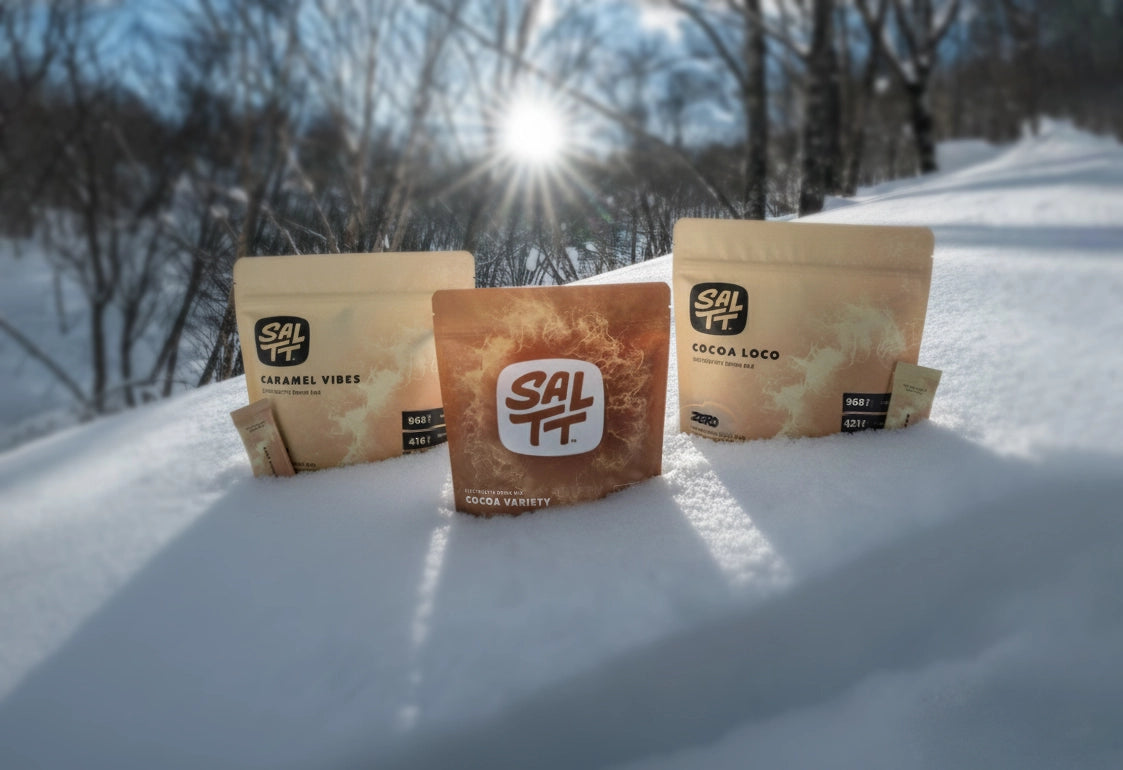 Three SALTT electrolyte drink mix pouches in snow: tan Caramel Vibes, brown Cocoa Variety, and tan Cocoa Loco. All feature water splash patterns. Cocoa Loco is Zero Sugars, Dyes, Citric Acid. Two small stick packs included.