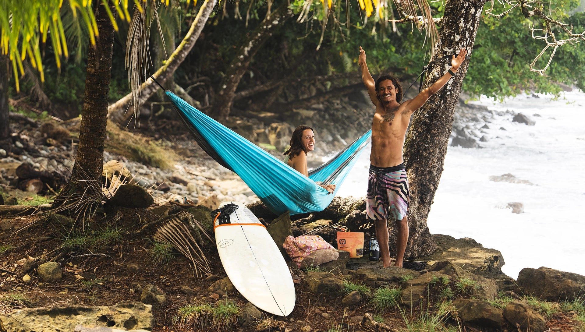 Two people relaxing in a hammock by a river with surfboards nearby.