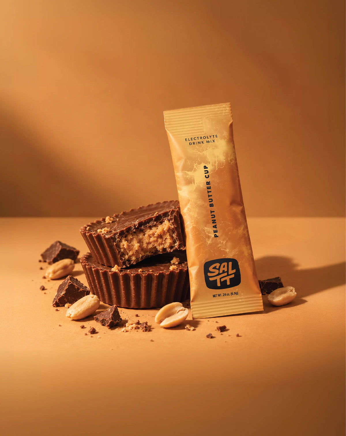 SALTT Peanut Butter Cup electrolyte drink mix stick pack with a tan ocean splash pattern beside chocolate peanut butter cups.