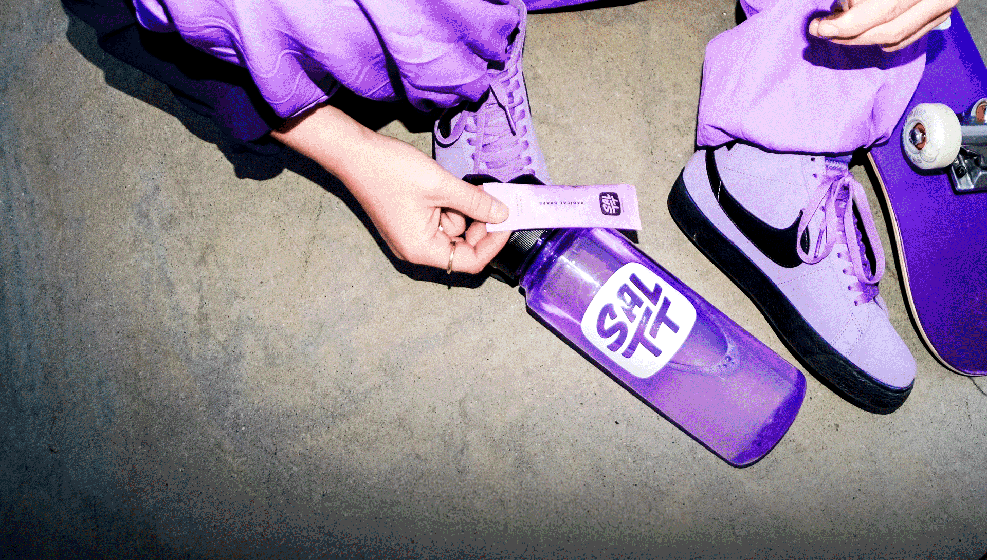 Purple SALTT Radical Grape Hydration Packet with ocean splash pattern held over a purple SALTT water bottle. Packet states ZERO Sugars, Dyes, Citric Acid.