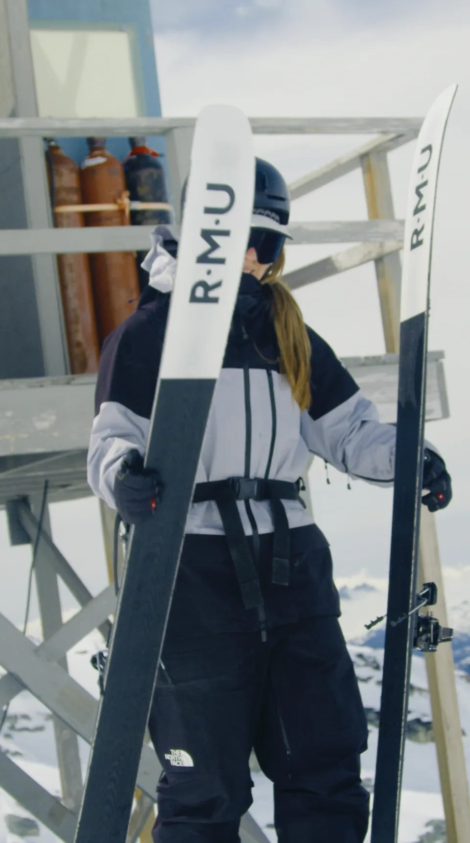 Skier wearing helmet, goggles holds R.M.U skis. North Face logo on black ski pants. Snowy mountain environment.