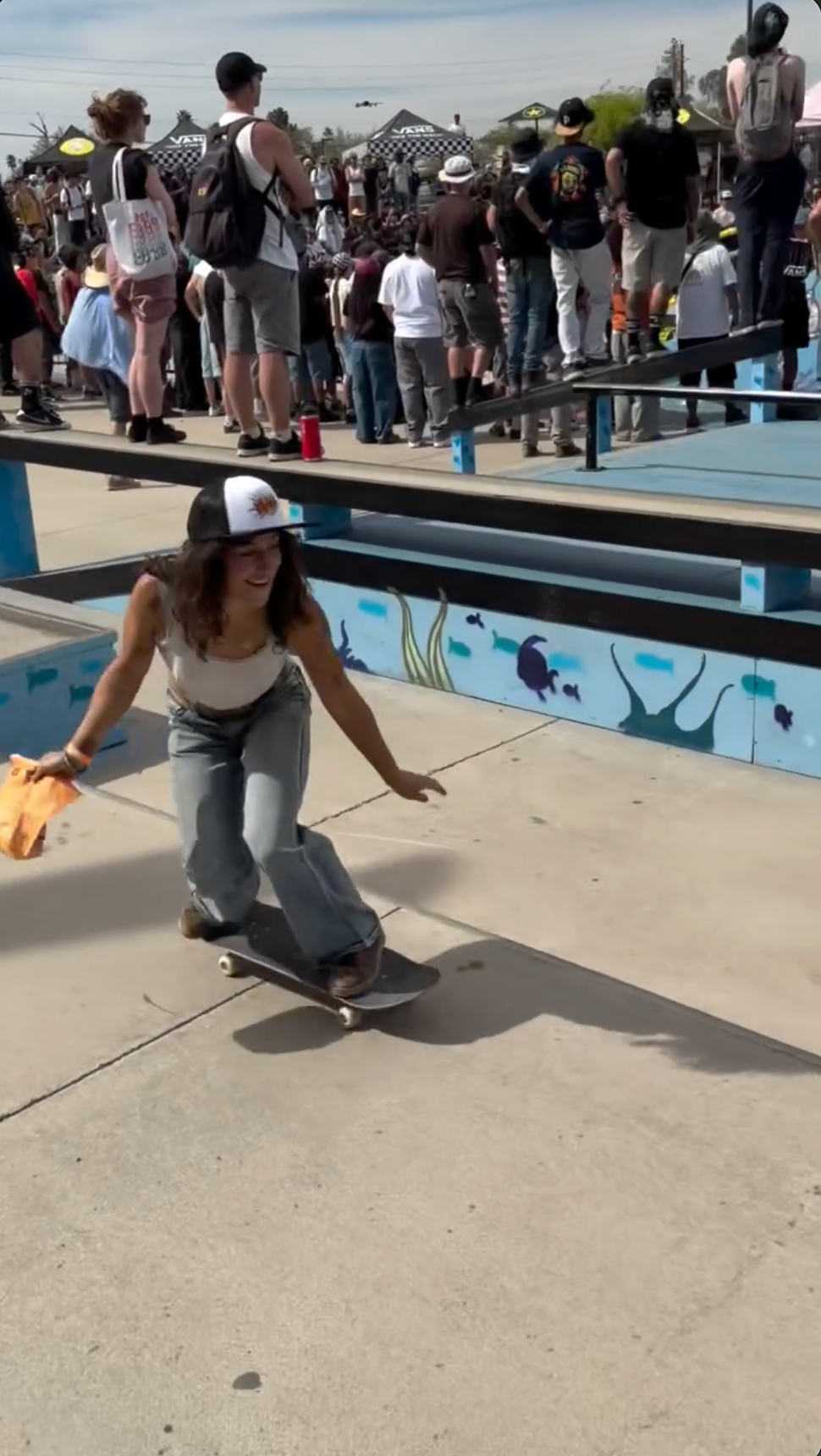 Smiling woman skateboards at a sunny park. Wears white top, jeans, cap, holds orange cloth. Blue walls with ocean murals and crowd watch.