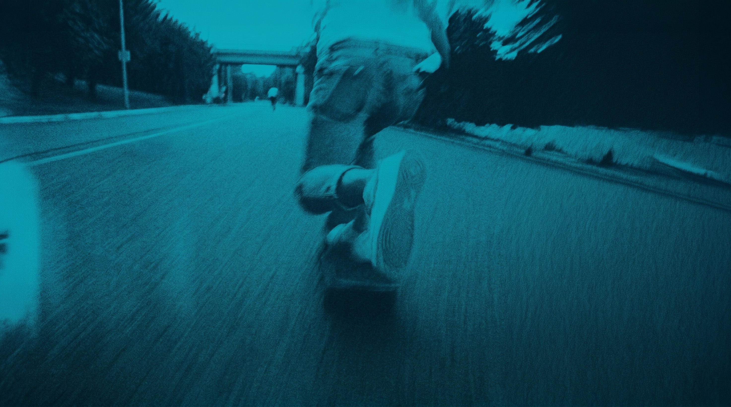 Low-angle, motion-blurred shot of a person skateboarding on an asphalt road, tinted in dark teal and blue-green, with trees and an overpass in the background.