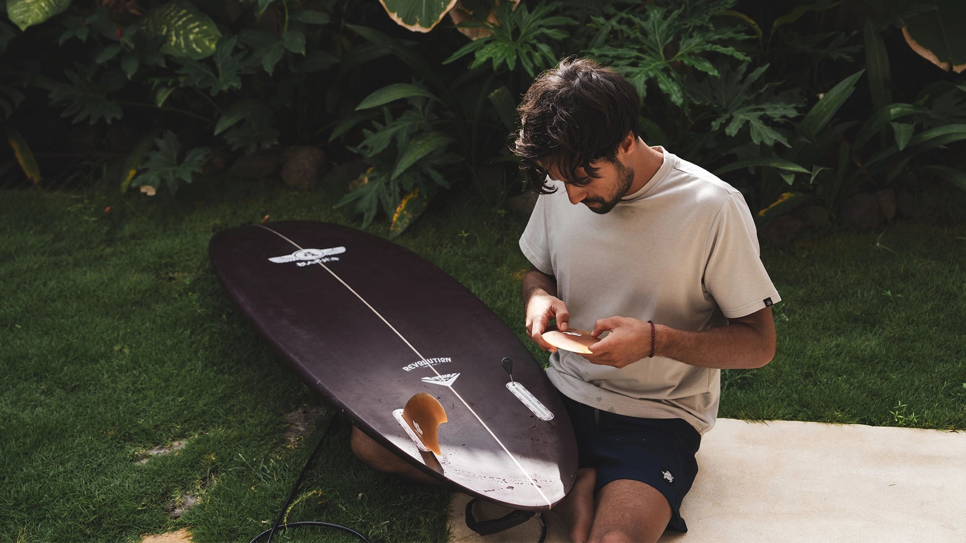 Man sitting on grass next to a surfboard with greenery in the background