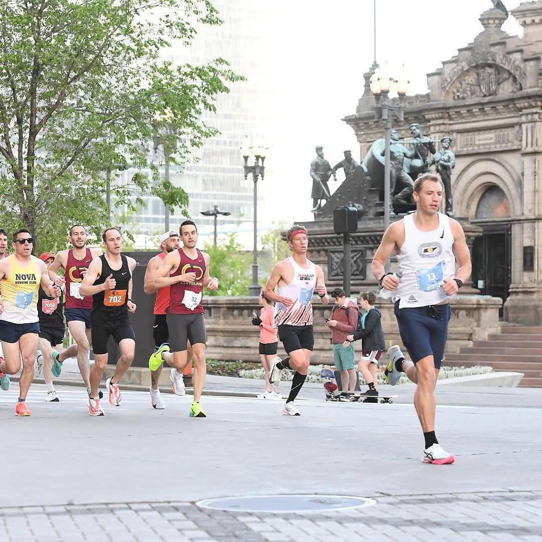 A group of male runners in athletic apparel. A runner in a yellow tank top displays NOVA RUNNING CLUB. Others wear maroon tank tops with a CLEVELAND logo. A runner in a white tank top features a Nike swoosh and bib number 13. Other visible bib numbers include 702, 9, 32, and 7050. Runners wear black, navy, and gray shorts, with running shoes in various colors including pink, neon yellow, and white. The background shows a city street with a large stone building and trees.