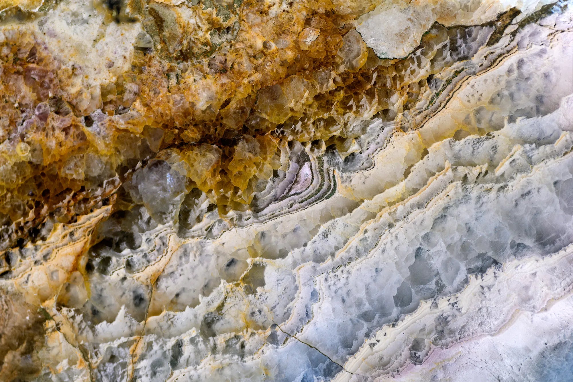 Close-up of a natural stone or mineral surface, featuring rough, layered textures with colors transitioning from golden yellow and orange on the left to white, gray, and light blue on the right, interspersed with darker bands and crystalline formations.
