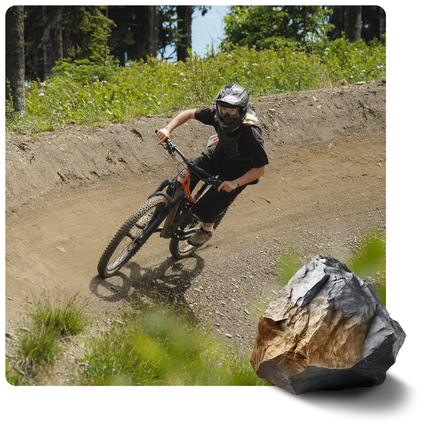A mountain biker in a helmet and goggles rides a black bike on a dirt trail. Green trees and foliage line the background. A large gray and brown rock is in the foreground.