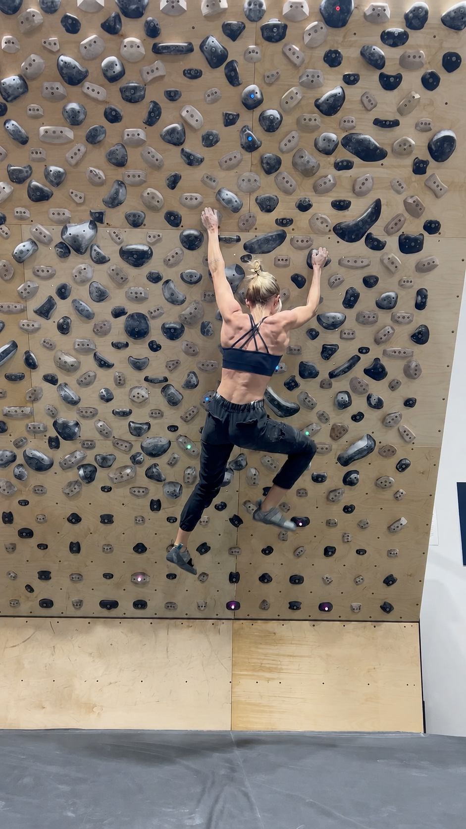 A woman in a black sports bra and dark pants climbs an overhanging wooden spray wall covered with rock climbing holds.