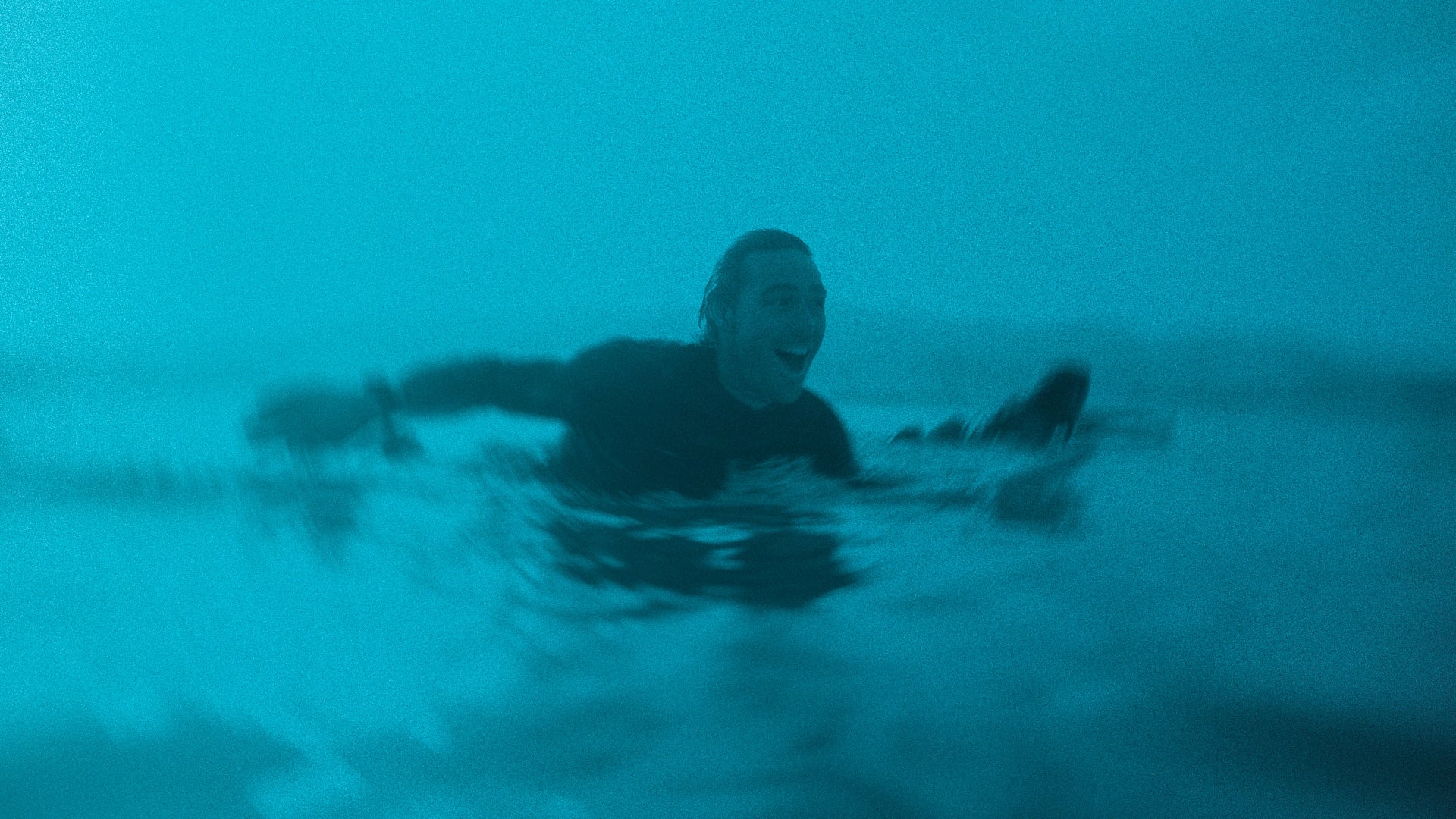 A person with a wide smile swims in blue-green water, with a surfboard partially visible beside them.