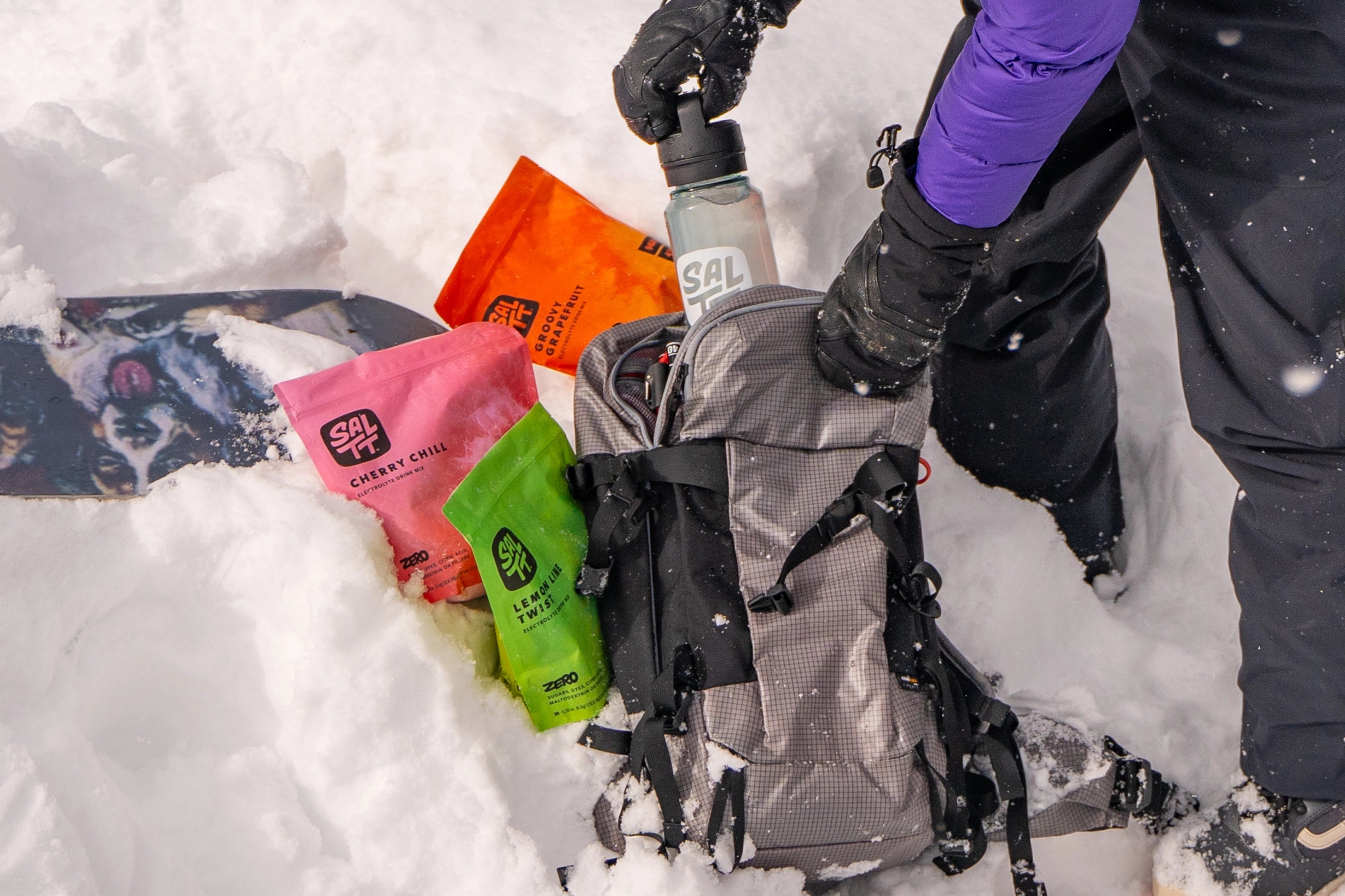 A snowy outdoor scene showing a backpack partially open with SALTT electrolyte drink mix pouches visible inside and nearby. The pouches include "Cherry Chill" (pink), "Groovy Grapefruit" (orange), and "Lemon Lime Twist" (green). A person in winter gear with a blue jacket and black gloves is placing a SALTT-branded water bottle into the backpack. A snowboard with a colorful graphic is partially buried in the snow.