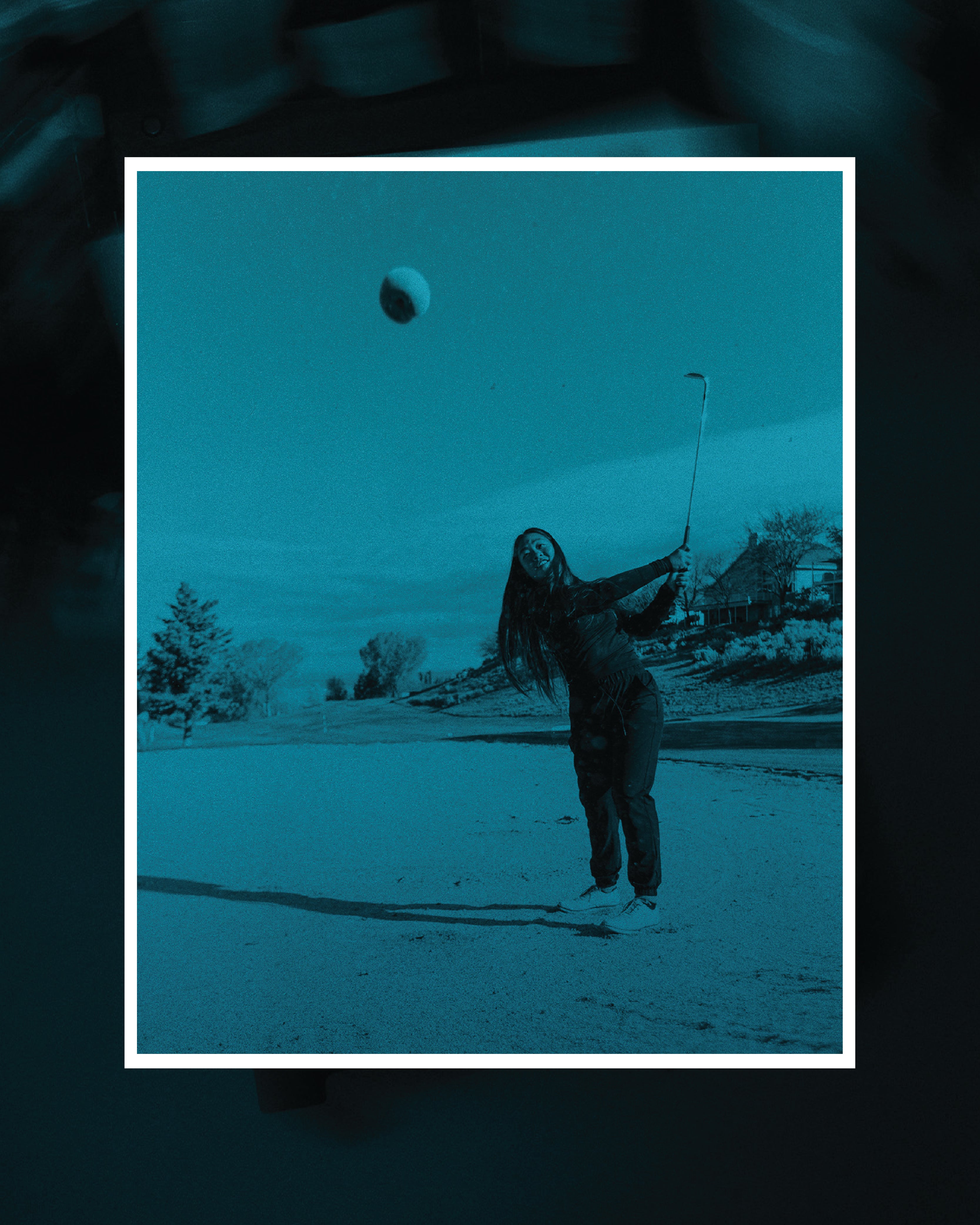 Teal-toned image of a woman in a golf bunker, mid-swing, with a golf ball airborne. Framed in white against a dark background.