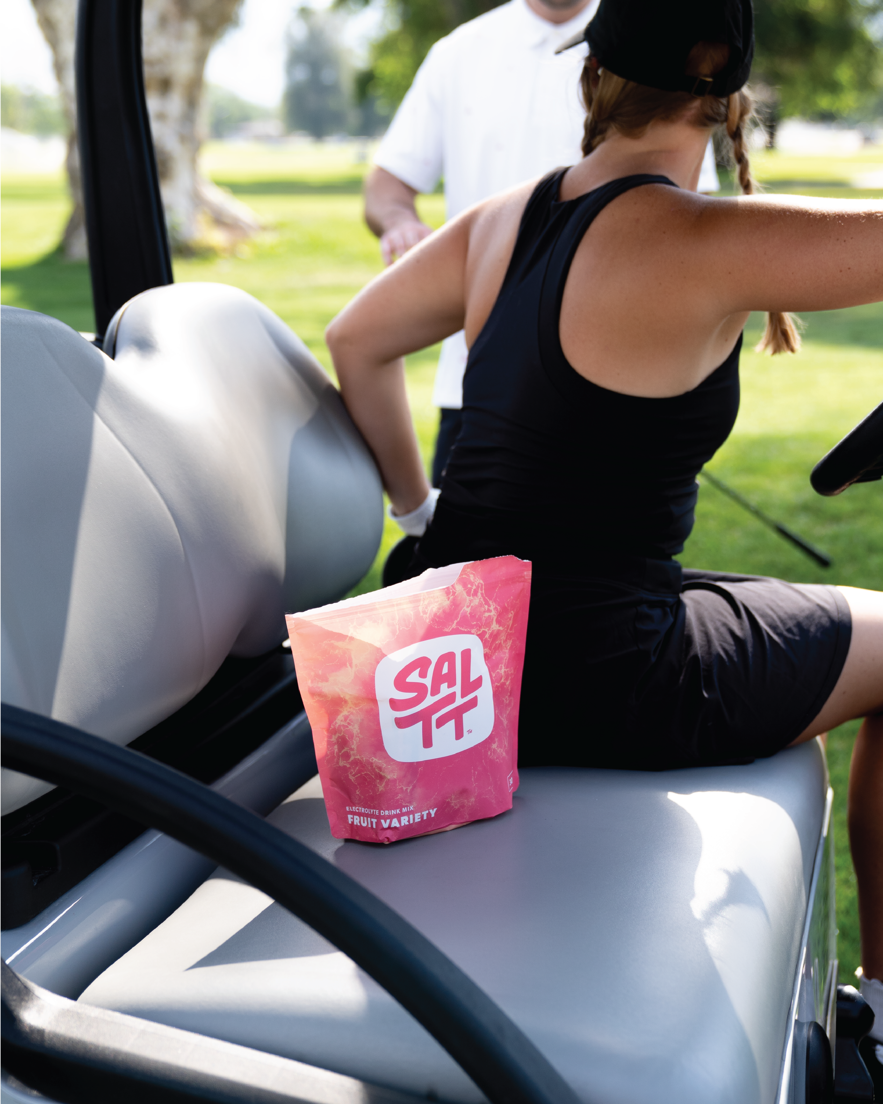 SALTT Fruit Variety Electrolyte Drink Mix pouch, colored pink with an ocean splash pattern and white text, resting on a golf cart seat.