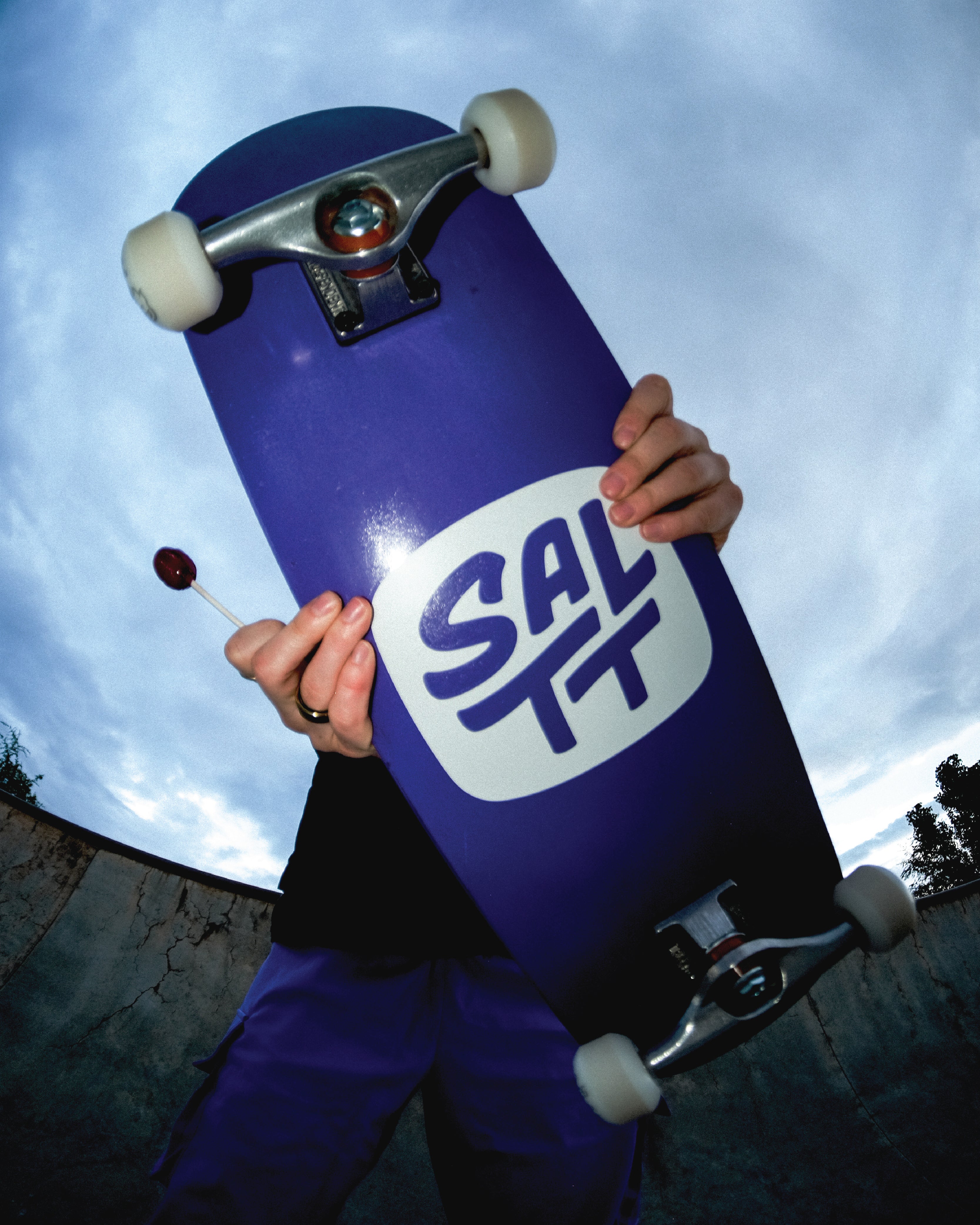 A person holds a purple skateboard featuring a white SALTT logo and a red lollipop, against a cloudy sky and concrete wall.