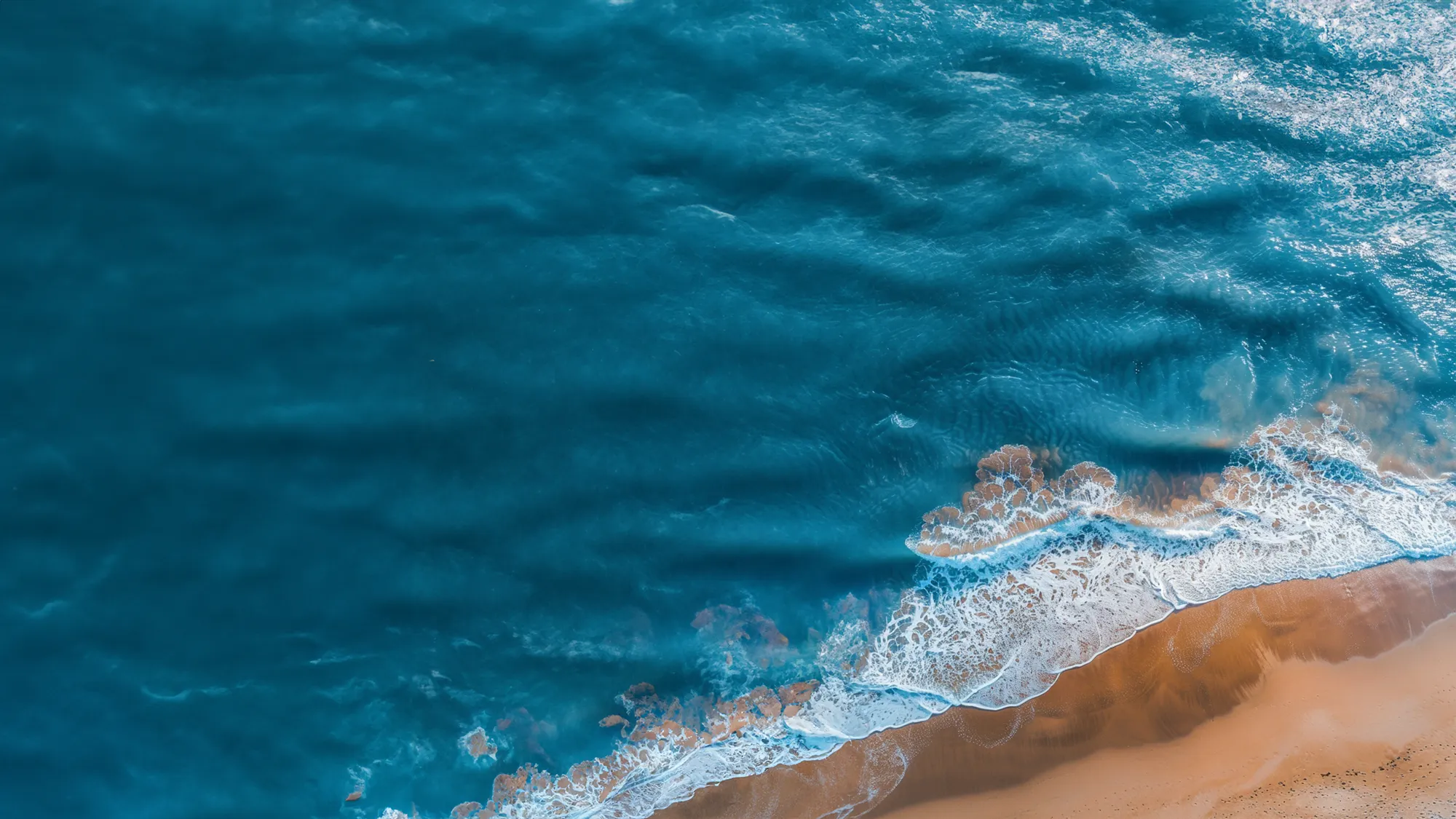 An aerial shot of a beach with blue waters rolling over sand.
