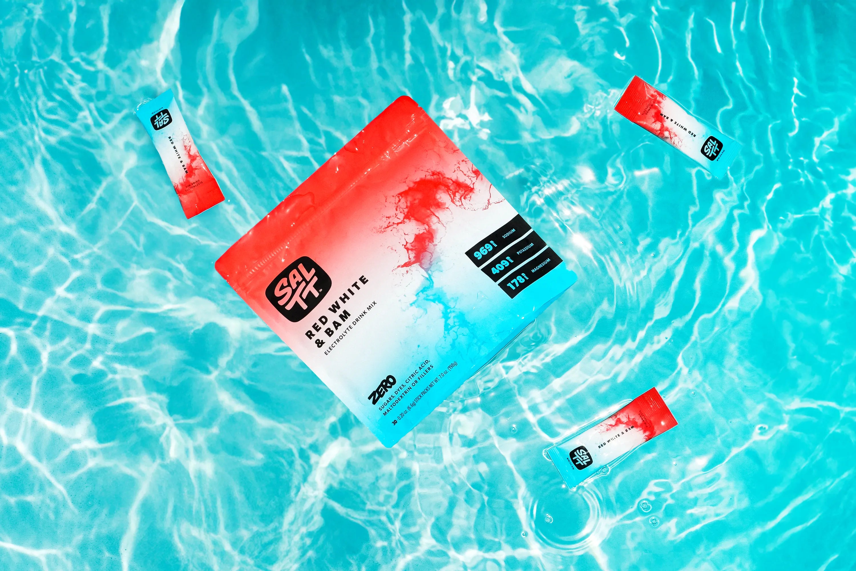 SALTT Red White & Bam Electrolyte Drink Mix large red, white, and blue ombre pouch with a red ocean splash design, displaying 969 Sodium, 409 Potassium, 178 Magnesium, and ZERO SUGARS, DYES, CITRIC ACID, MALTODEXTRIN OR FILLERS, floats in blue water beside three smaller matching stick packs.