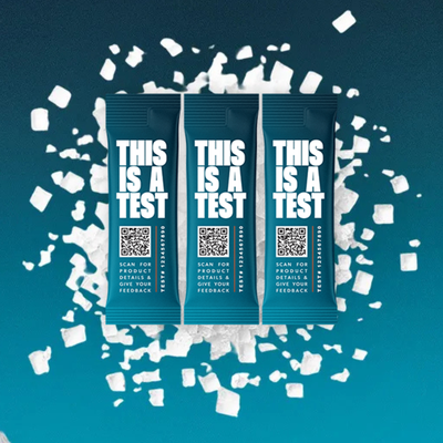 Three dark teal SALTT THIS IS A TEST single-serve stick packs on a teal and dark blue background with scattered white salt crystals.