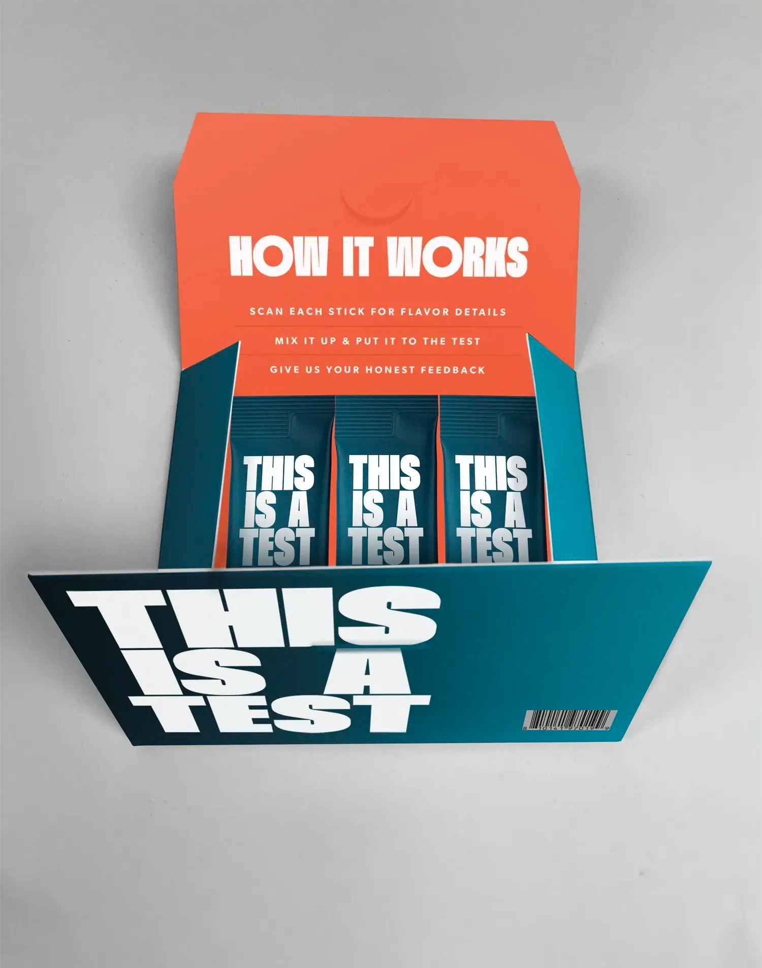 Open product packaging box with a dark teal base and an orange inner flap. It contains three dark teal stick packs. The box front and each individual stick pack display large white text THIS IS A TEST. The orange inner flap features white text HOW IT WORKS and multiple lines of usage instructions. A barcode is visible on the front of the box.