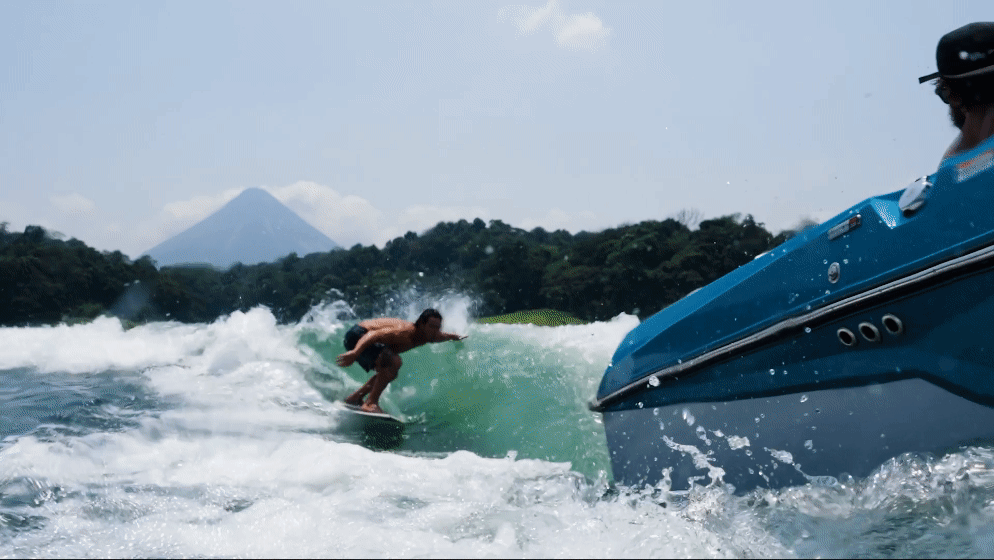 A person on a board rides a green wake behind a blue and grey boat. Green trees and a grey volcano are visible in the background under a light blue sky.