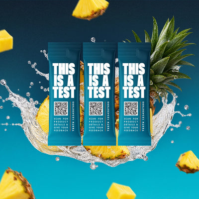 Three blue single-serve test packets labeled THIS IS A TEST surrounded by pineapple slices and water splashes on a blue background.