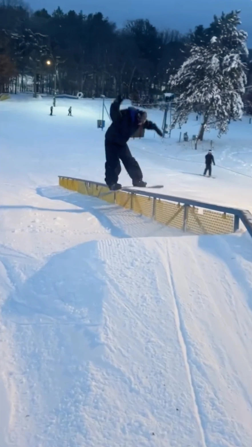 A snowboarder in dark clothing slides sideways on a yellow rail. The snowy slope has trees and other people in the background under a dark blue sky.