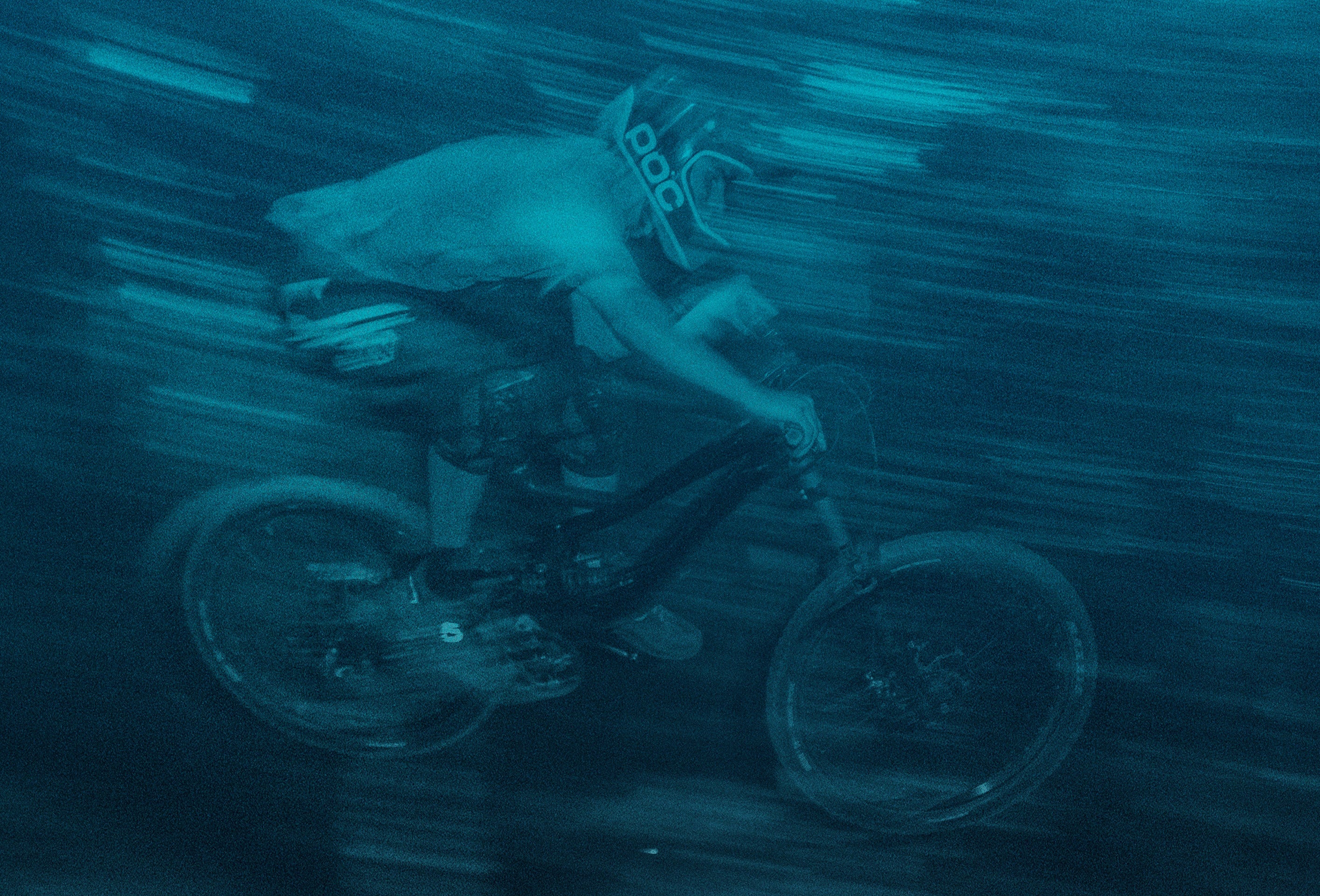 A mountain biker wearing a helmet with a POC logo, blurred by motion, against a dark teal streaky background.