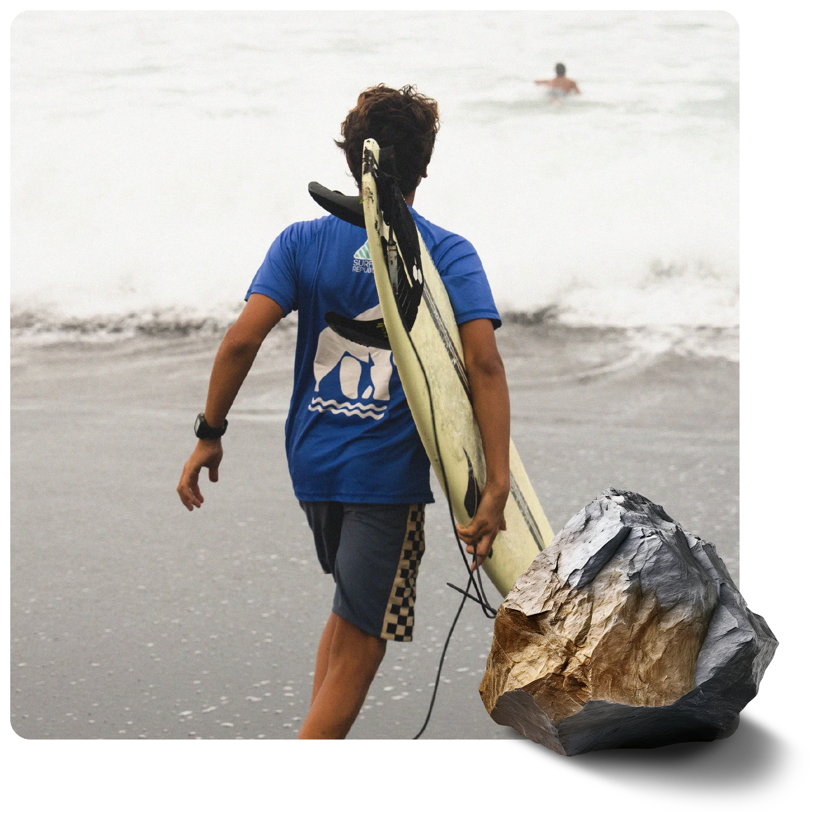 A person with a surfboard walks into the ocean, waves breaking. Another person swims in the distance. A textured gray and gold rock is in the foreground.