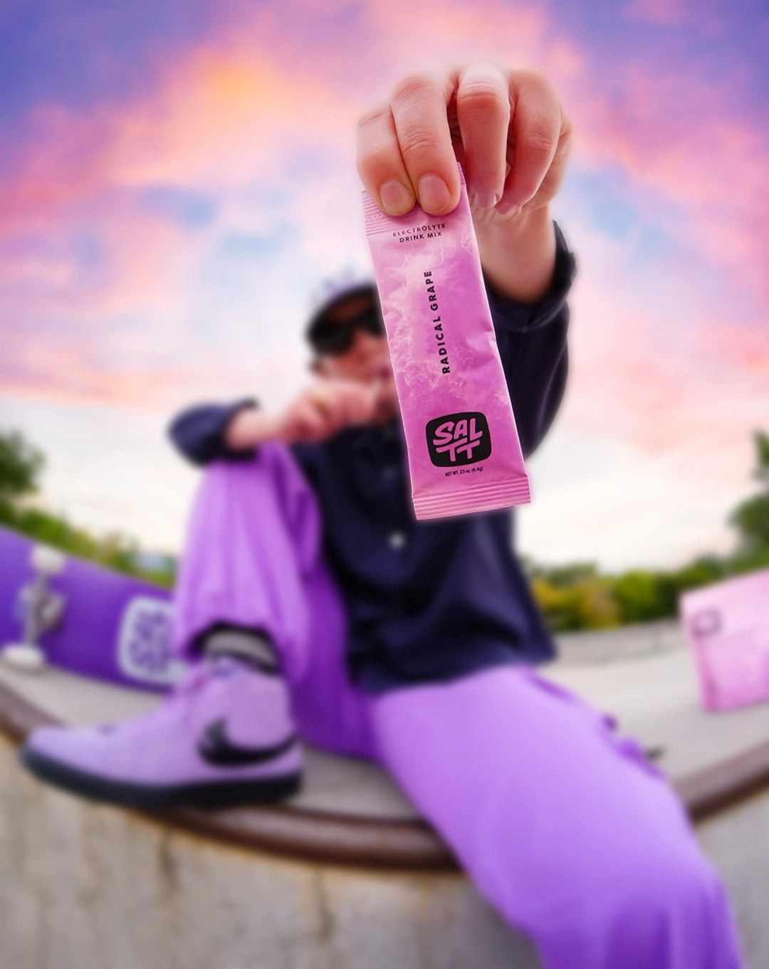 A hand holds a pink SALTT Radical Grape Electrolyte Drink Mix single-serve stick pack with an ocean splash pattern.