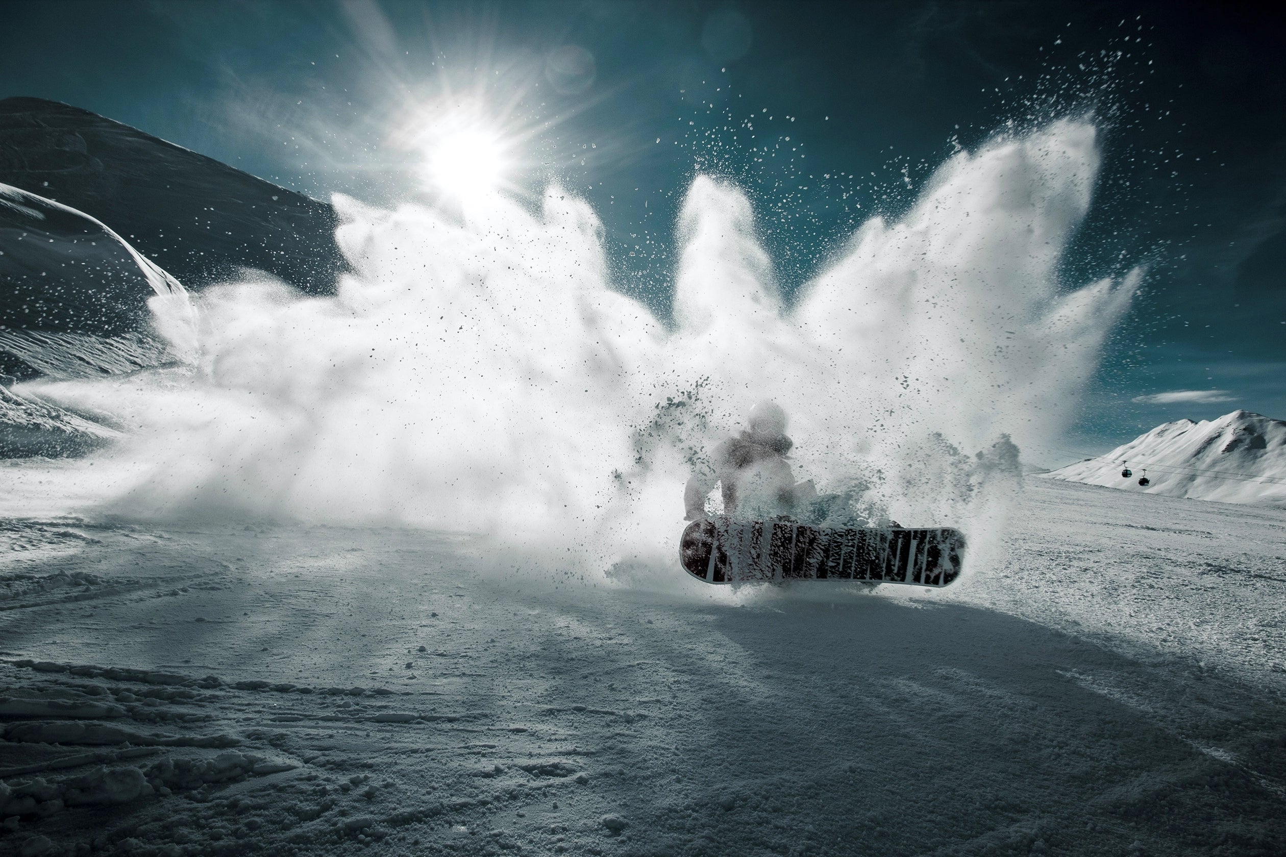 A snowboarder creates a large spray of white snow on a dark colored snowboard with white markings on a snowy mountain, under a bright sun in a dark blue sky.
