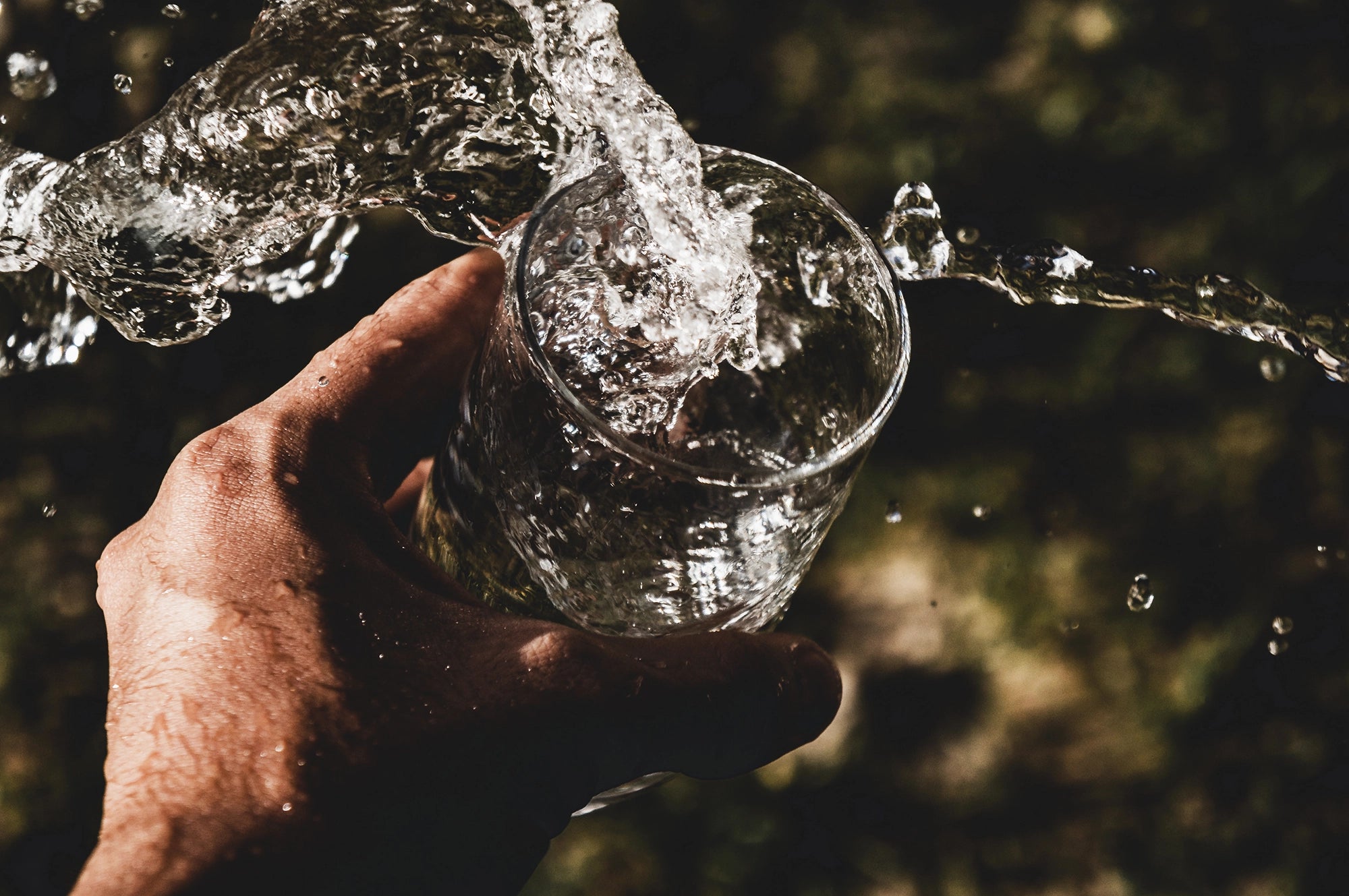 A person's hand holds a clear glass as a stream of water pours into it, creating splashes. The background is dark and out of focus.