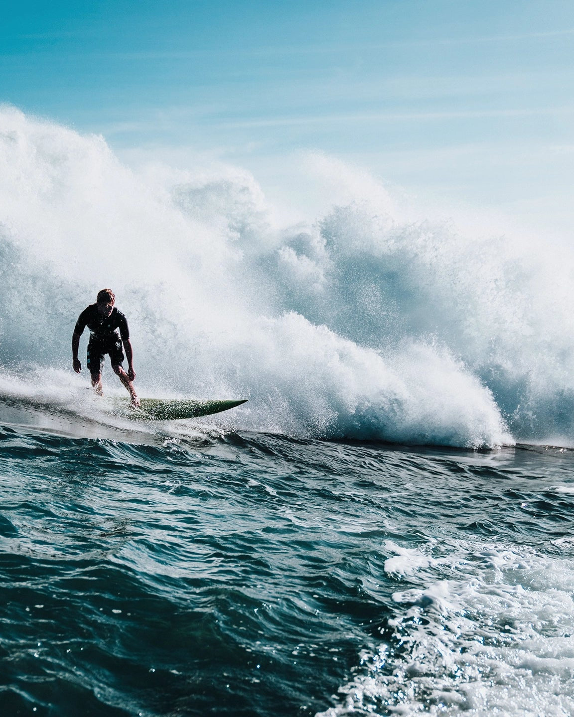 A surfer in a dark wetsuit rides a green surfboard on a large, powerful ocean wave. White water crashes behind the surfer, with deep blue water in the foreground and a light blue sky above.